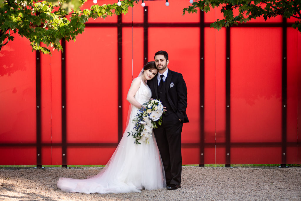 Bride and groom stand close together in front of a red wall